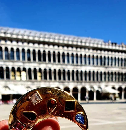 Gold-tone 1980s Yves Saint Laurent crescent brooch set with coloured stones, held up against the arcades of a Venetian square