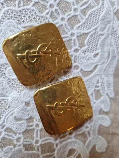 Pair of gold-tone 1980s Yves Saint Laurent square clip earrings with the monogram, lying on lace