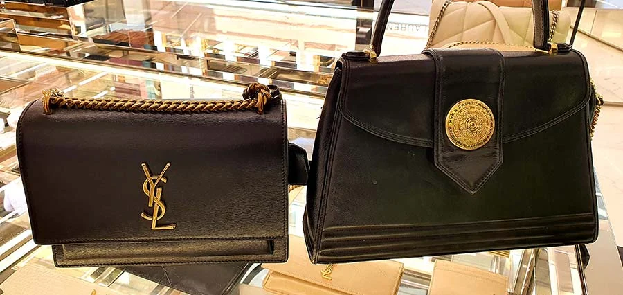 Black Yves Saint Laurent 2021 bag with a gold logo beside a vintage 1980s black bag with a round gold medallion, in a shop