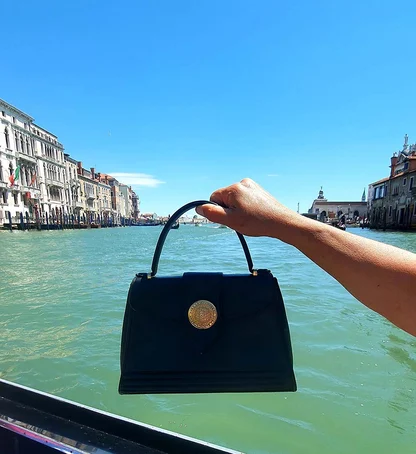 Black vintage Yves Saint Laurent handbag with a gold medallion clasp, held up over the water in Venice