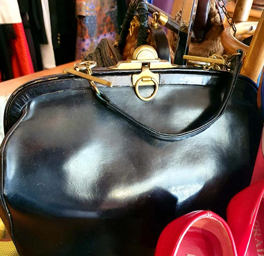 Roberta di Camerino black leather handbag with a brass clasp and ring fastening, on a market stall beside red shoes