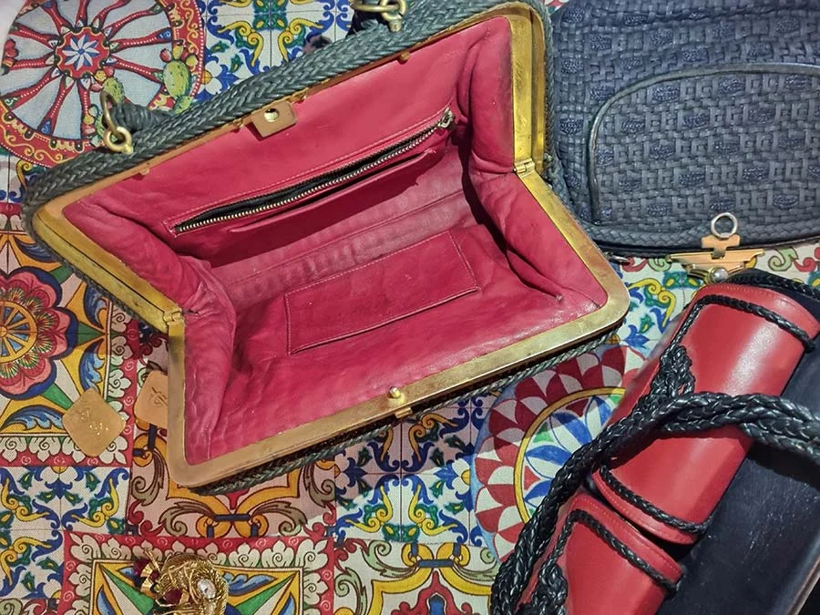 Open woven bag with a gold frame and plush red lining, beside a navy woven bag and a red and black leather one