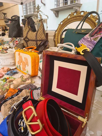 Market stall display with a wooden box, red leather belts with gold buckles, a woven box bag and an ornate gold mirror