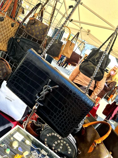 Black crocodile-effect leather bag with a chain strap and metal monogram, among handbags on a market stall