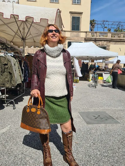 A smiling woman in a burgundy leather jacket and brown lace-up boots holding a monogram Louis Vuitton bag at a market