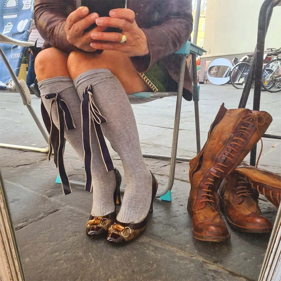 Grey knee socks tied with black ribbons, worn with buckled heels, beside a pair of tan lace-up leather boots