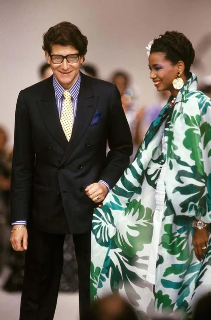 A man in glasses and a dark double-breasted suit beside a woman in a green and white leaf-print cape, at a fashion show