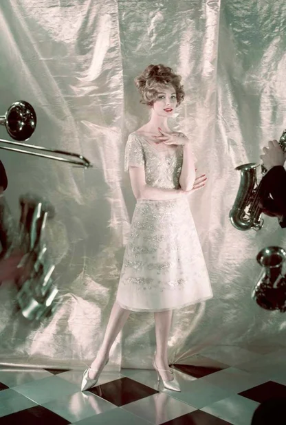 Woman in a pale beaded knee-length dress posing on a chequered floor among brass instruments