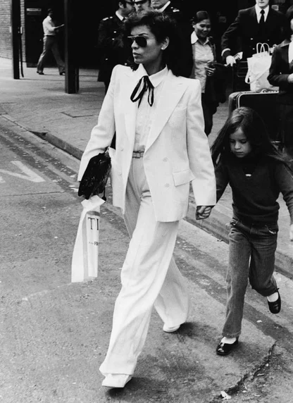 Black-and-white street shot of a woman in a white trouser suit with a black bow tie and sunglasses, holding a girl's hand