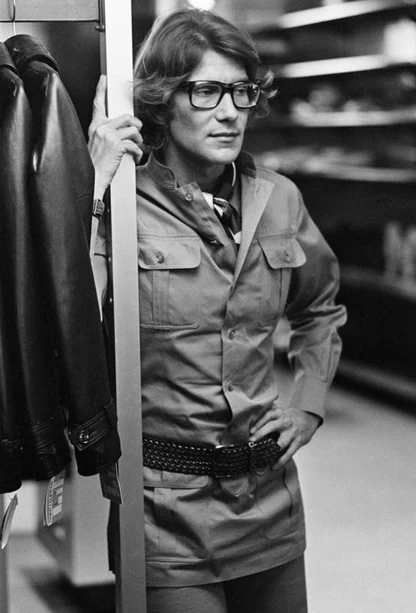 Man in glasses and a safari-style shirt jacket with a woven belt, leaning on a shop rail beside a leather jacket
