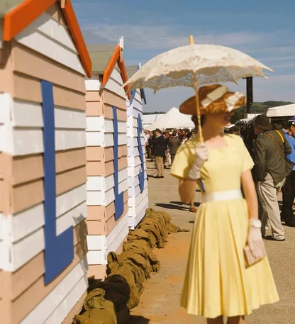 Woman in a yellow 1950s-style dress, straw hat and white gloves holding a lace parasol beside striped beach huts