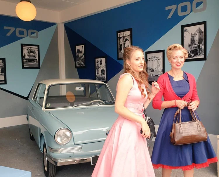 A woman in a pink sleeveless dress and another in a navy dress with a red cardigan, beside a pale blue vintage car