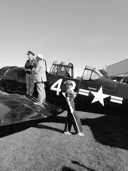 Woman in a cap, sailor-style top and flared trousers beside a vintage propeller plane while two men stand on its wing