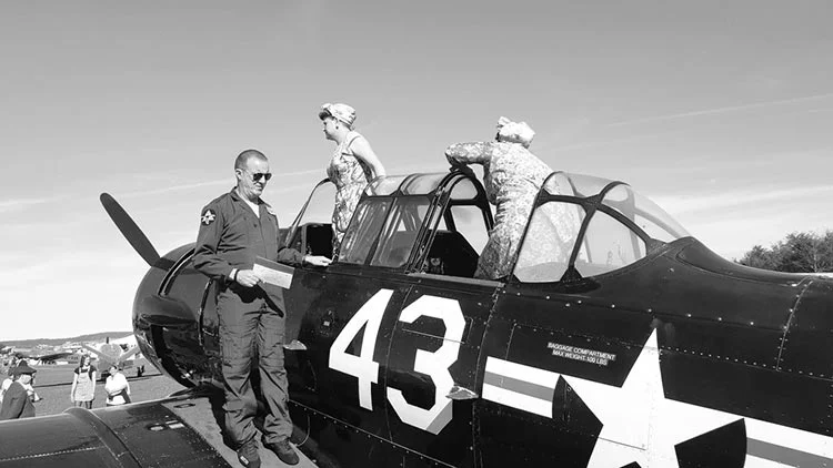 Black-and-white shot of two women in 1940s-style outfits and a man in a flying jacket on a propeller plane marked 43