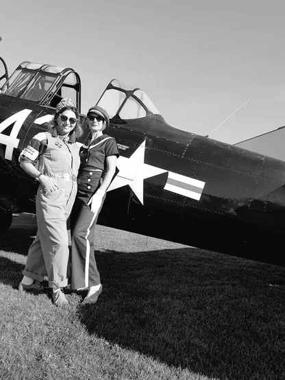 Two women in vintage-style overalls and a sailor-style outfit standing in front of a black propeller plane with a white star