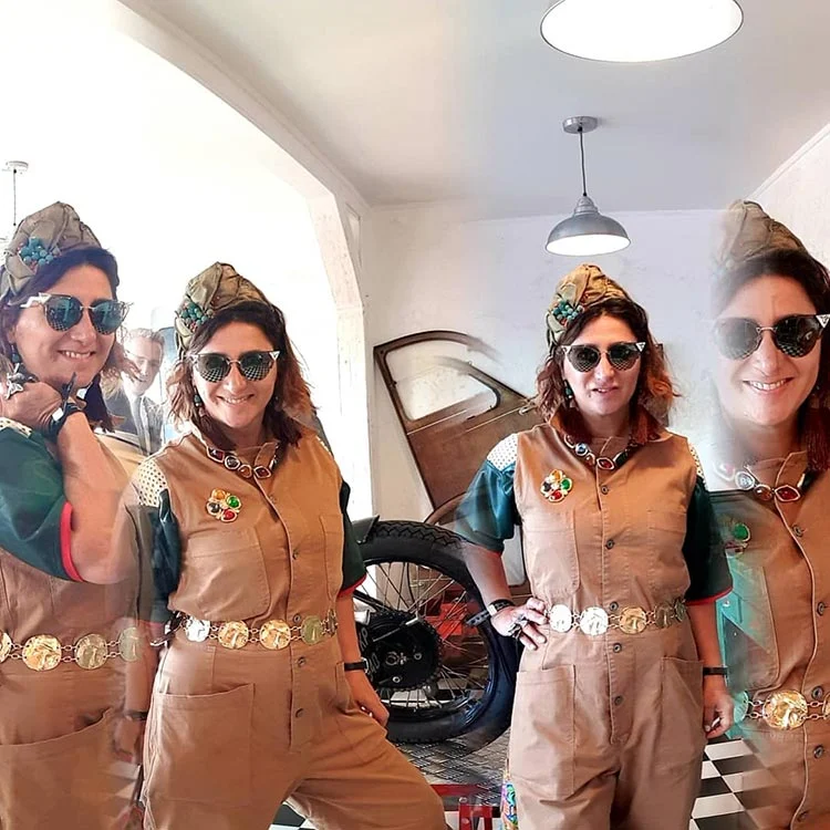 Gracie Opulanza in tan overalls with a gold chain belt, khaki forage cap and sunglasses, in front of a vintage motorbike