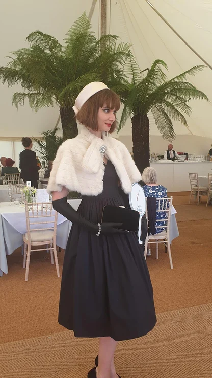 Woman in a black full-skirted dress, white fur stole, pillbox hat and long black gloves, holding a clutch in a marquee