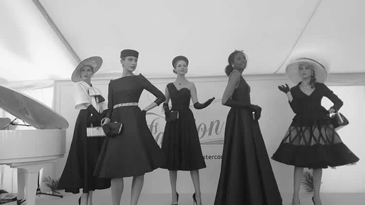 Five models in dark 1950s-style dresses, hats and gloves on the Dior vintage catwalk, a white piano at the left