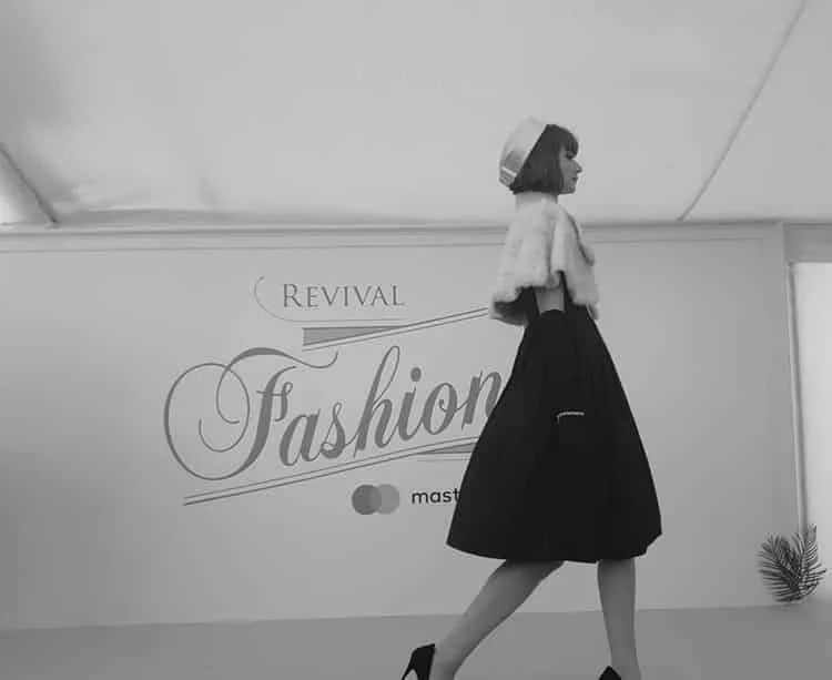 Model in a black full-skirted dress and white fur stole walking past the Revival Fashion sign at the Dior catwalk