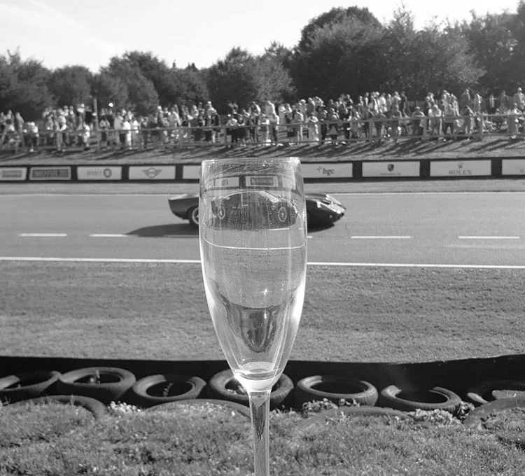 Black-and-white view through a champagne flute of a vintage racing car passing spectators at Goodwood Revival
