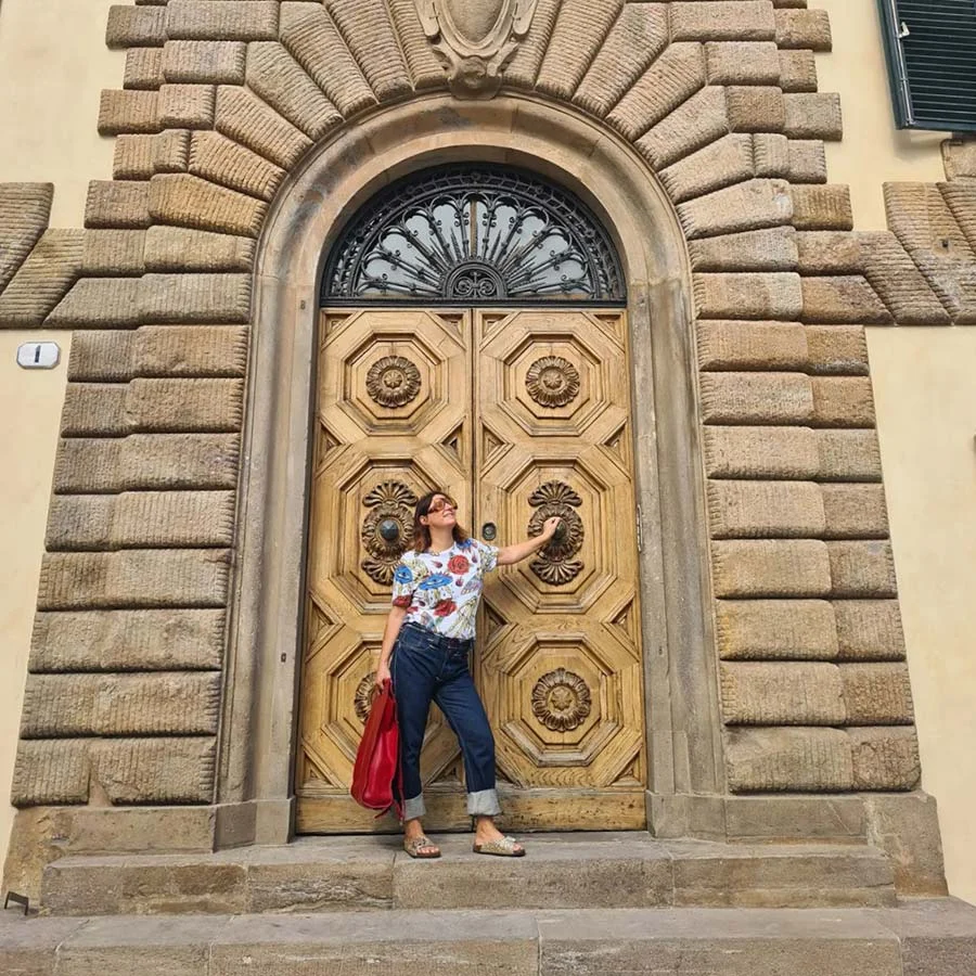 Gracie Opulanza in a floral top and Levi's reaching towards a large panelled wooden door framed by rough stonework