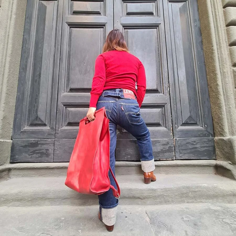 Gracie Opulanza in a red jumper, cuffed Levi's and tan boots, holding a red tote at a tall grey door