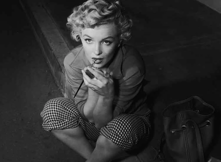 Marilyn Monroe sitting on a kerb in gingham capri trousers, putting on lipstick, with a bag beside her