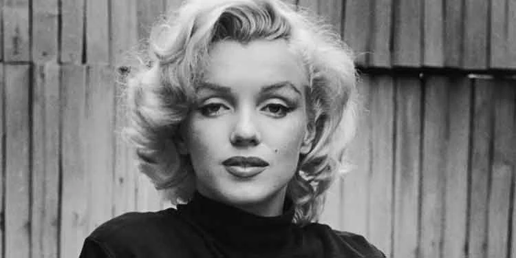 Close black-and-white portrait of Marilyn Monroe in a black polo neck against a wooden fence