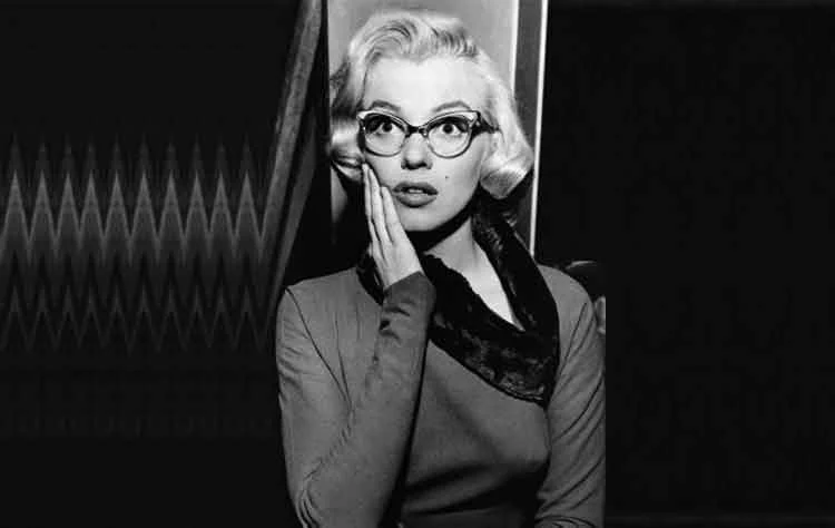 Marilyn Monroe in cat-eye glasses and a dark top with a lace collar, one hand to her cheek