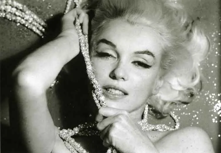 Marilyn Monroe lying back with a rope of diamanté necklaces held at her cheek, in a black-and-white photograph