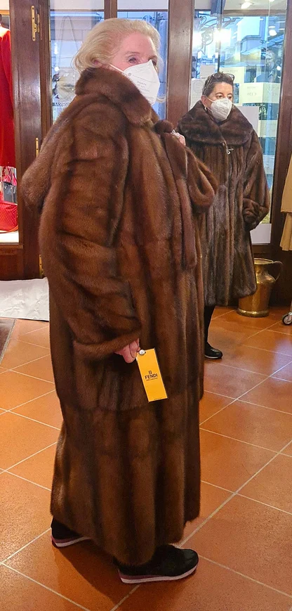 A woman in a long brown mink Fendi coat with the Fendi tag on its pocket, in a shop, another fur coat behind her