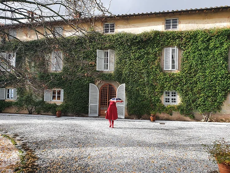 Woman in a red cape holding an umbrella on a gravel courtyard in front of an ivy-covered Italian villa