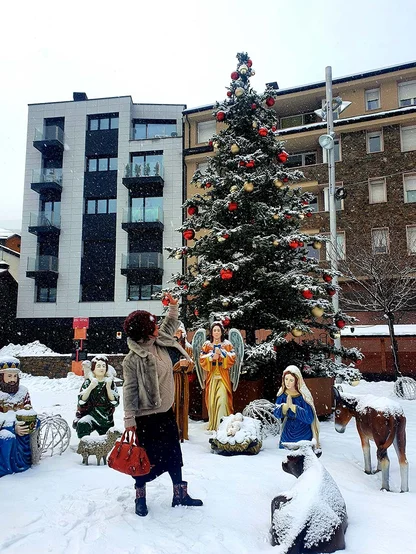 A woman in a dark hat and grey jacket holding a red bag in a snowy square, beside a Christmas tree and nativity figures