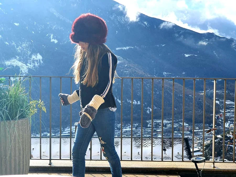 Girl in a burgundy faux fur hat, knitted mittens and jeans on a terrace with a snowy mountain behind