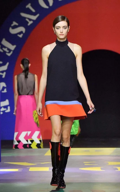 Model in a navy halterneck dress with a blue and orange hem and black boots, against a red and blue runway backdrop