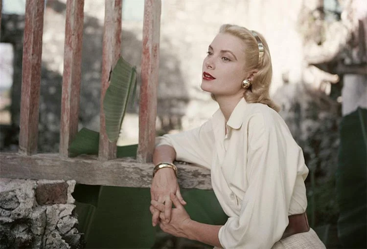 Grace Kelly in a cream blouse, gold bracelet and headband, leaning on a wooden fence with her gaze to one side