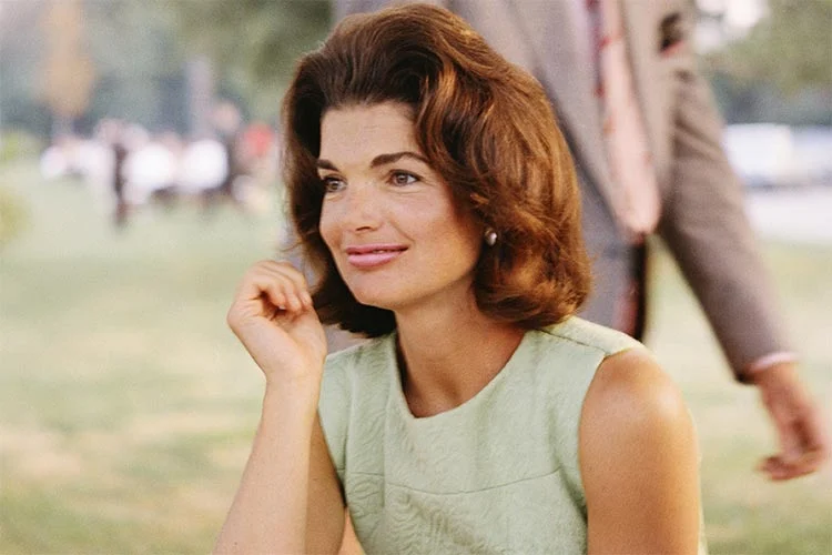 Jackie Kennedy in a pale green sleeveless dress with a pearl earring and a bouffant, her chin resting on one hand