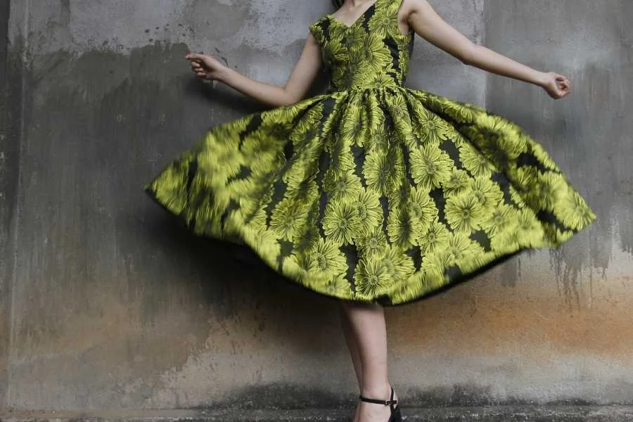 Full-skirted green dress with a black floral pattern, swirling as the wearer turns, against a stained grey wall