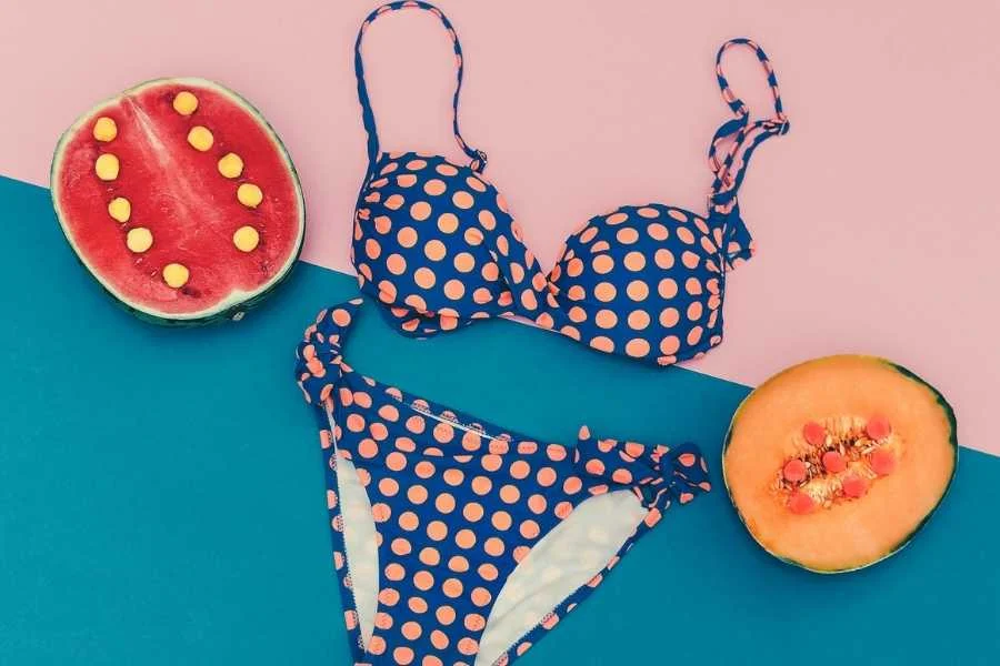 Navy bikini with orange polka dots laid flat on a pink and teal background, beside slices of watermelon and melon