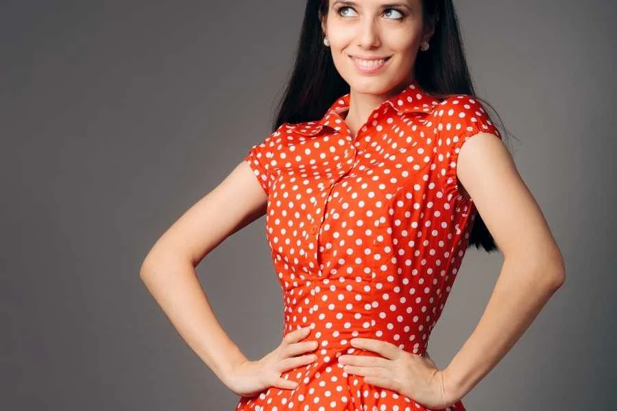 Smiling woman with long dark hair in a red polka dot dress with cap sleeves and a collar, hands on hips