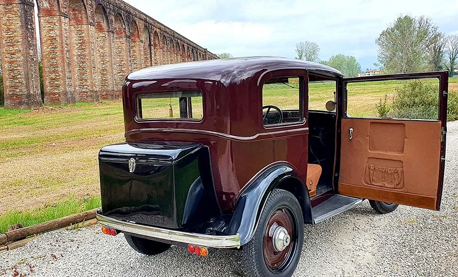 Maroon and black Fiat Balilla with its door open, parked in Tuscany beside a long stone aqueduct