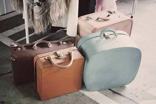 Four vintage suitcases in brown, tan, pale blue and pink standing on a pavement beside a clothes rail