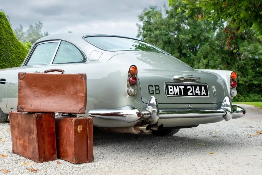 Silver Aston Martin seen from the rear, with three worn tan leather suitcases stacked beside the bumper