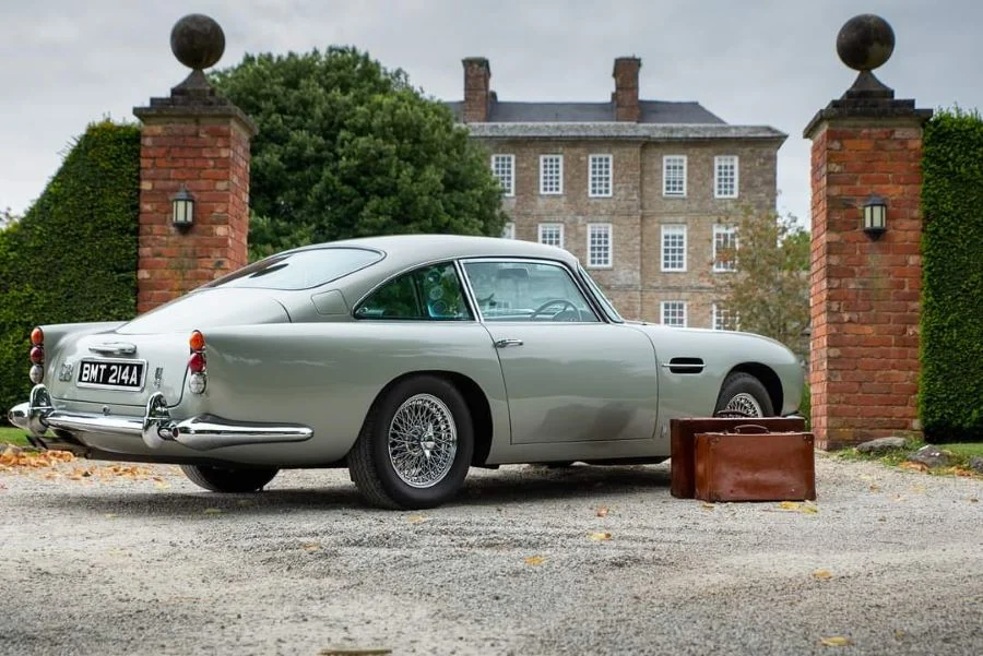 Silver Aston Martin on a gravel drive in front of a brick country house, two leather suitcases by the rear wheel