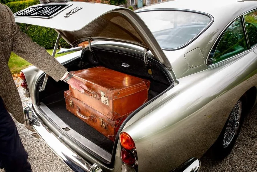 Open boot of a silver Aston Martin holding two tan leather vintage suitcases