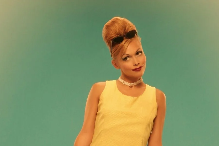 Woman with a tall beehive hairstyle, sunglasses pushed into her hair, a pearl choker and a yellow sleeveless top
