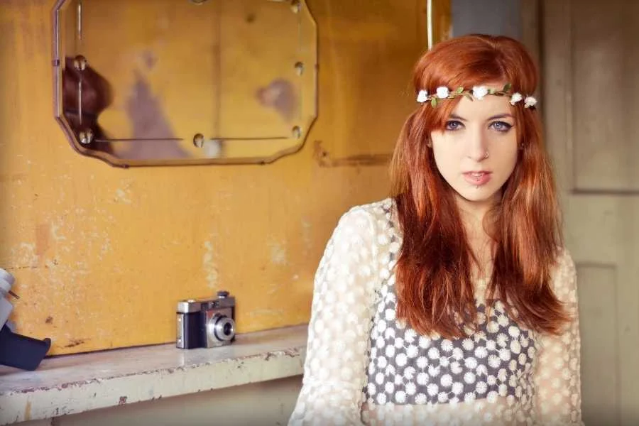 Young woman with long red hair and a flower headband, in a lace-print top beside a vintage camera on a shelf