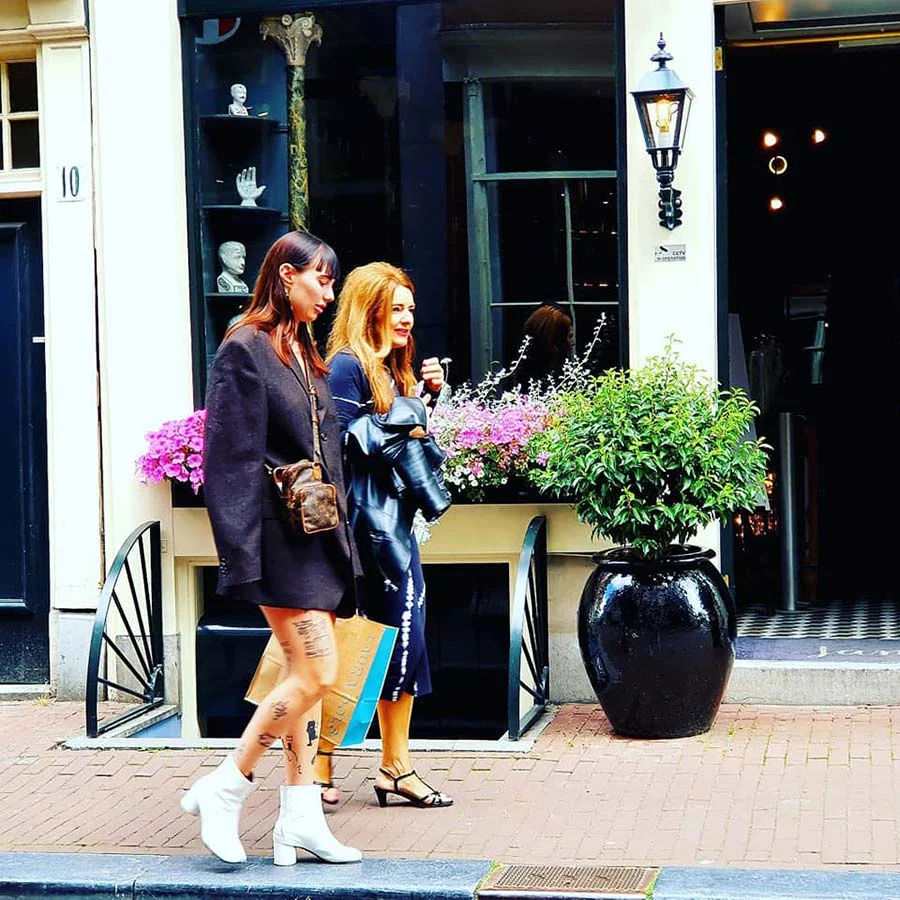 Two women walking past a shop window in Amsterdam, one in a dark blazer and white ankle boots with a small tan shoulder bag