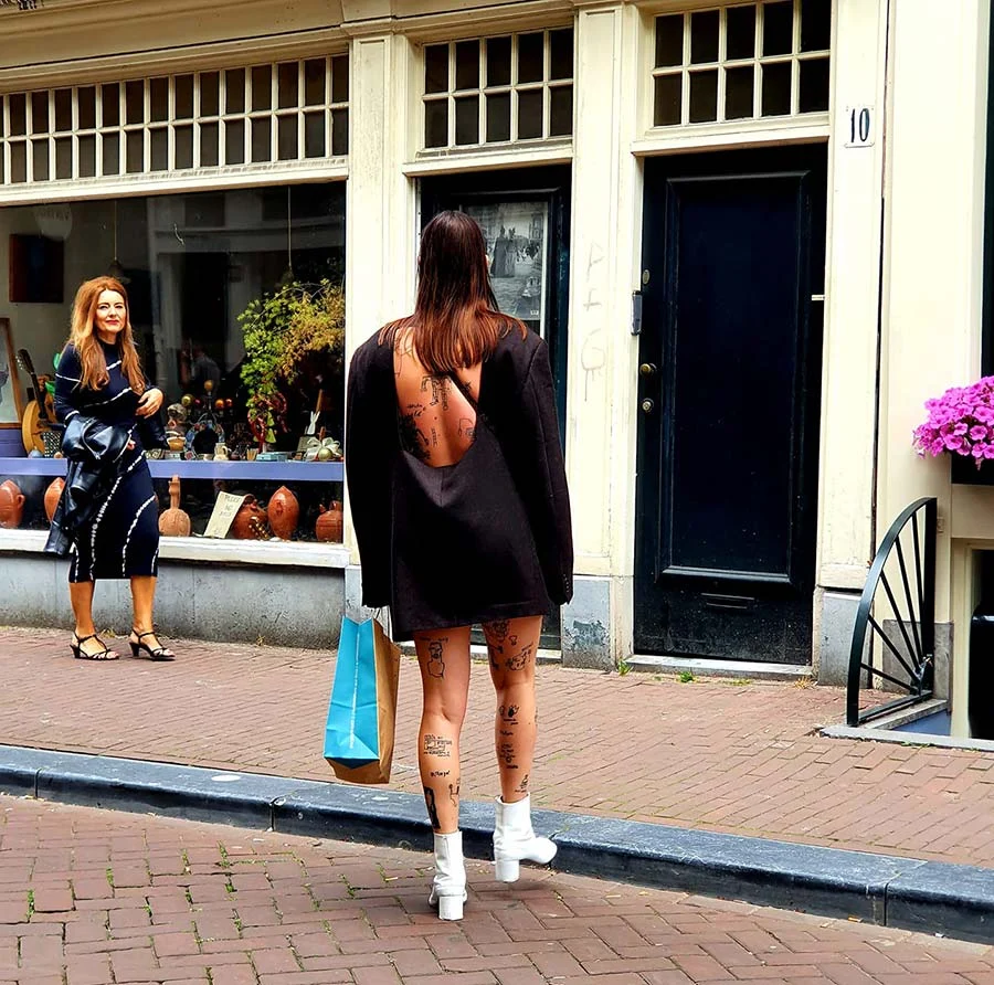 Woman on an Amsterdam street in an oversized black blazer worn backless, with white ankle boots and a blue shopping bag