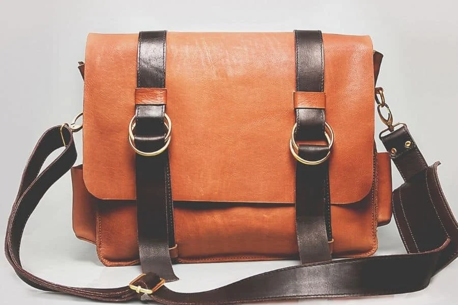 Tan leather satchel with two dark brown straps and brass rings, with a long shoulder strap
